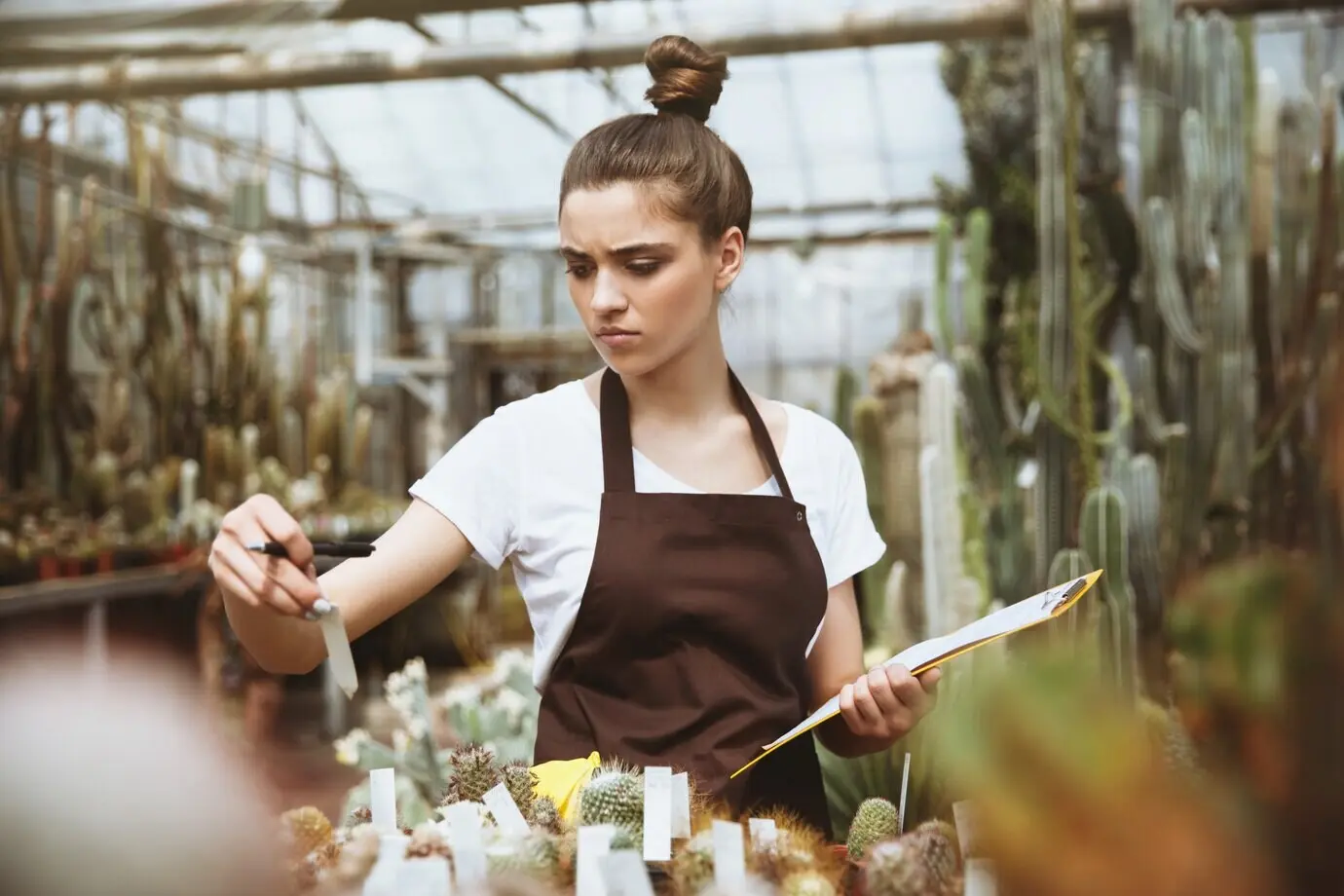Ernste junge Frau steht in einem Gewächshaus und hält ein Klemmbrett.